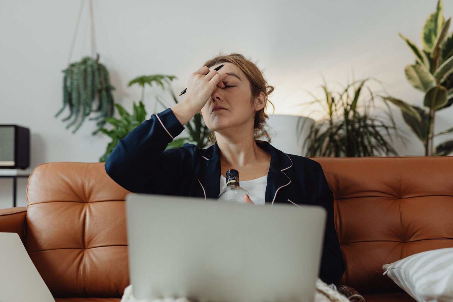 Woman sitting on a couch with a laptop, holding a liquor bottle and rubbing her forehead.