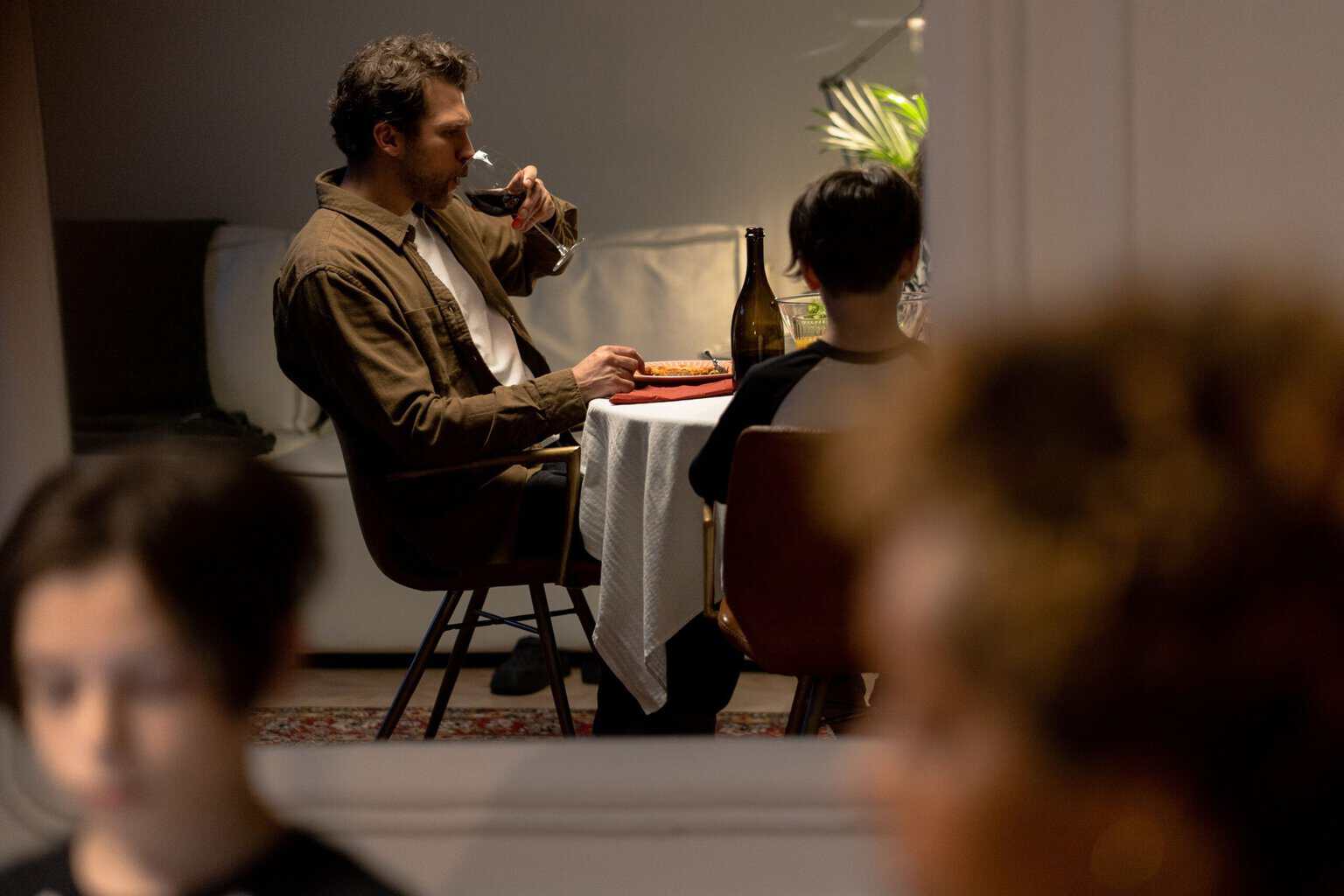 Man drinking a glass of red wine alone at the dinner table while a child and other family members sit nearby, illustrating heavy drinking and the impact of alcohol misuse on family relationships.
