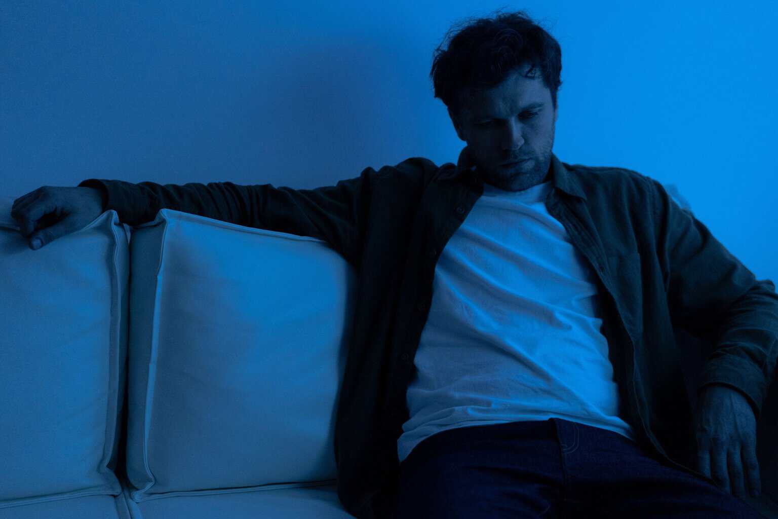 Young man sitting slumped on a white couch in a dimly lit room with blue lighting, looking down with a withdrawn expression, representing the emotional effects of heavy drinking and alcohol misuse.
