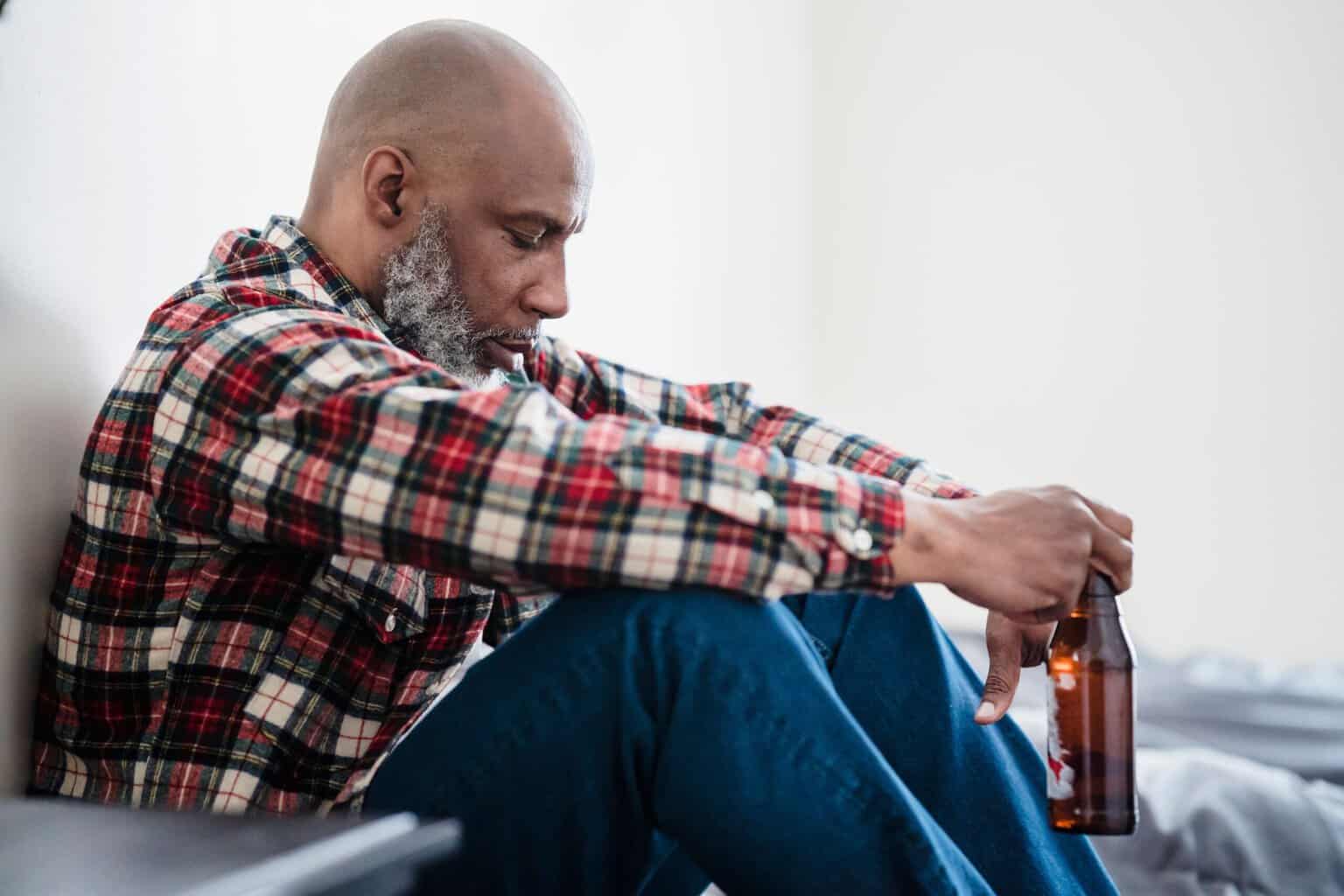 Man sitting alone on the floor holding a beer bottle, representing the isolation and emotional impact of alcoholism and alcohol addiction.