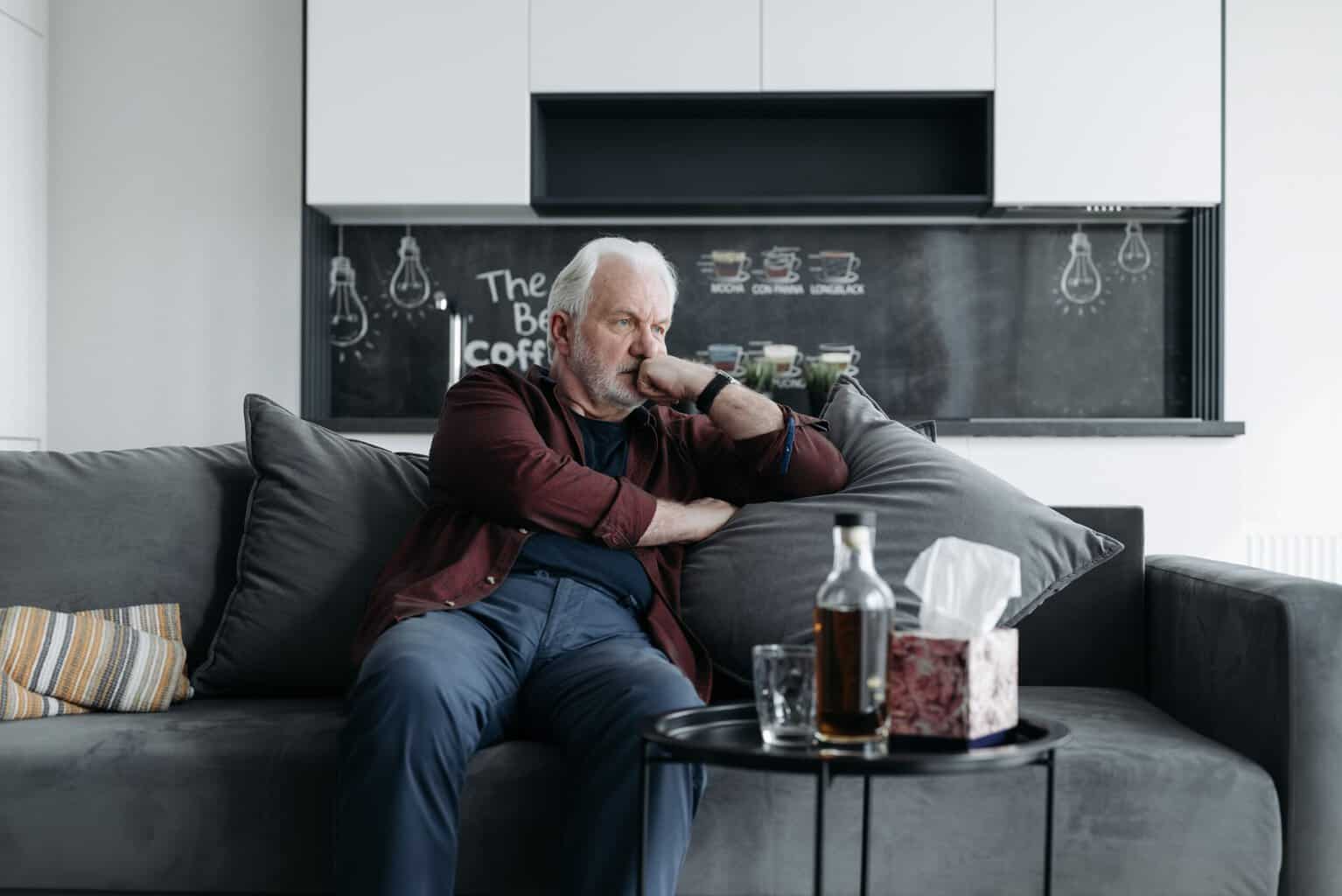 Senior man sitting thoughtfully on a couch with a bottle of alcohol and an empty glass on a nearby table, illustrating alcohol dependence.