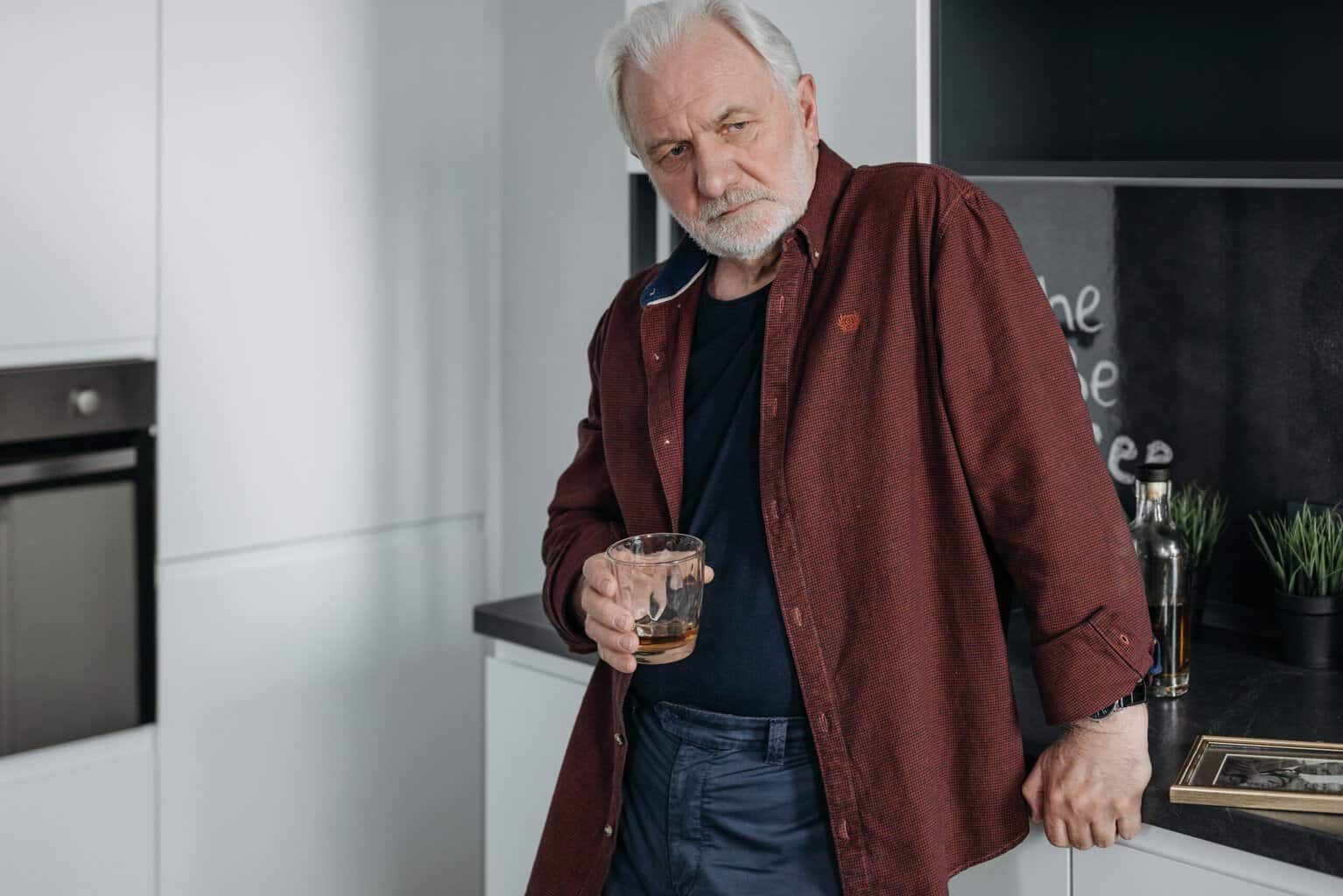Older man standing in a kitchen holding a glass of whiskey, representing alcohol dependence, alcoholism, and the challenges of alcohol addiction.