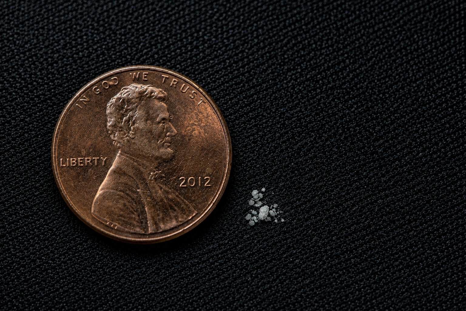 Close-up of a U.S. penny next to a tiny amount of white powder on a dark textured surface to illustrate the small amount of fentanyl that can be dangerous.