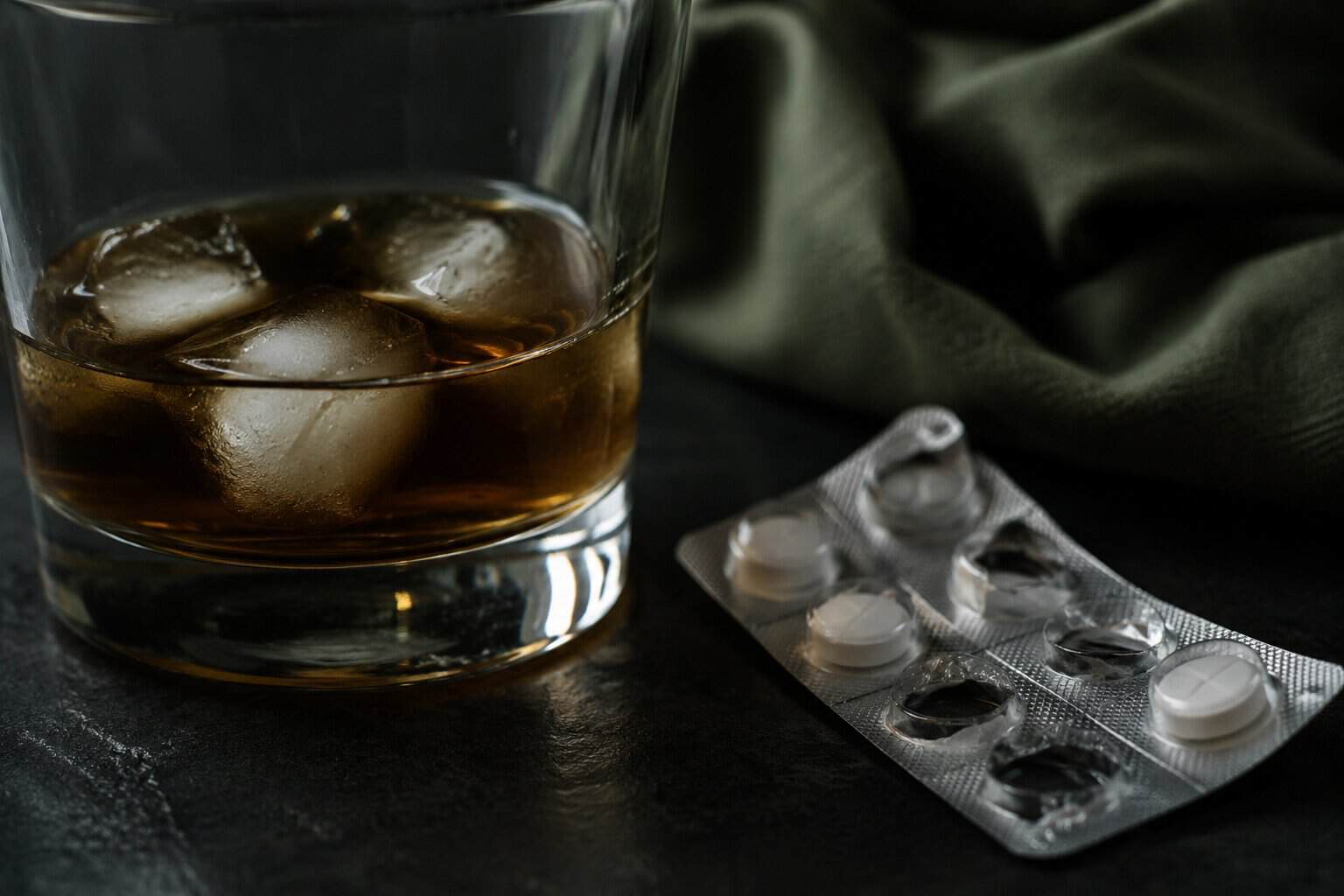 Close up of a glass of alcohol beside a blister pack of white antidepressant pills on a dark surface for an article about mixing alcohol and antidepressants.