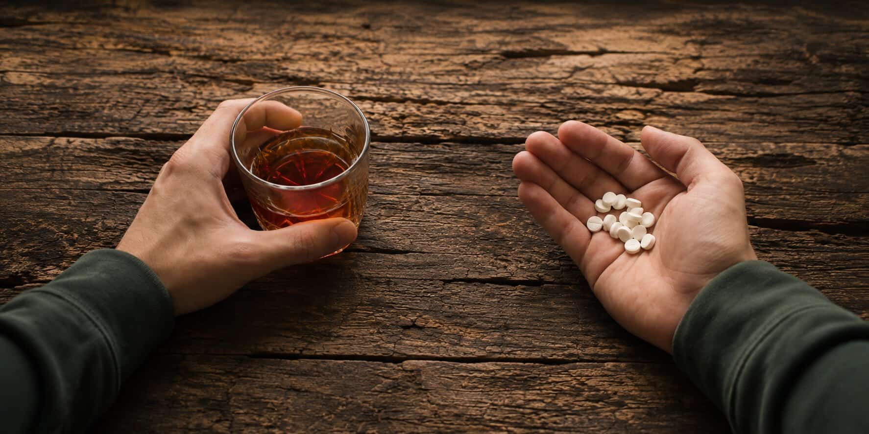 Overhead view of a person holding a glass of alcohol in one hand and white antidepressant pills in the other for an article about mixing alcohol and antidepressants.