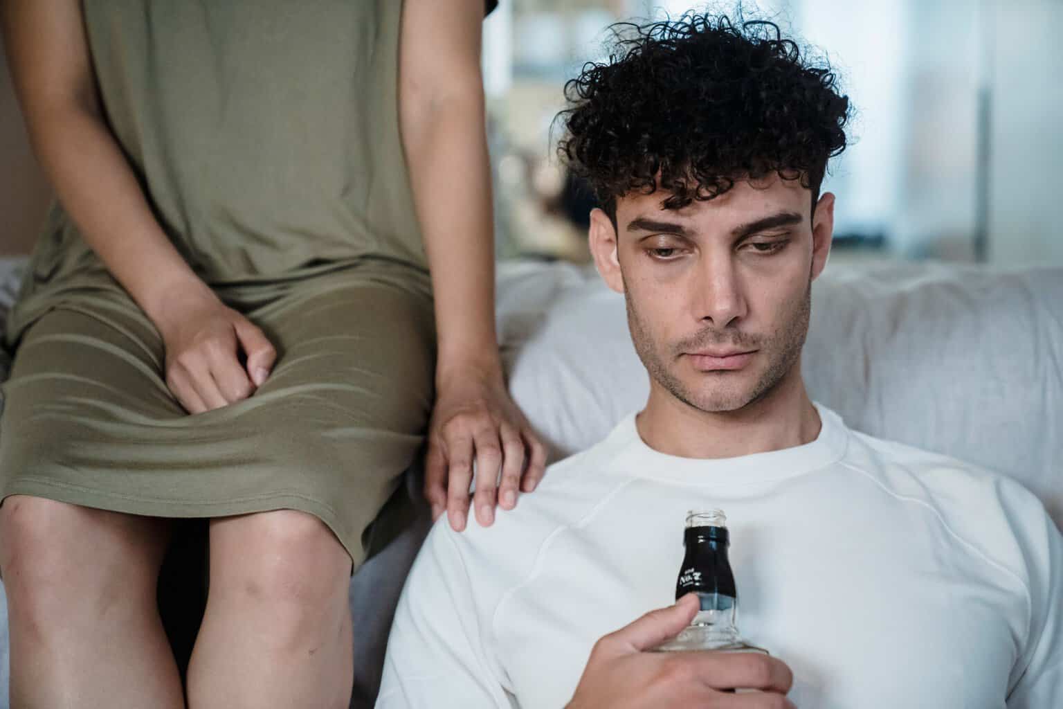 Man holding a bottle of alcohol while sitting beside his partner, who rests a supportive hand on his shoulder, representing the impact of problem drinking on relationships.